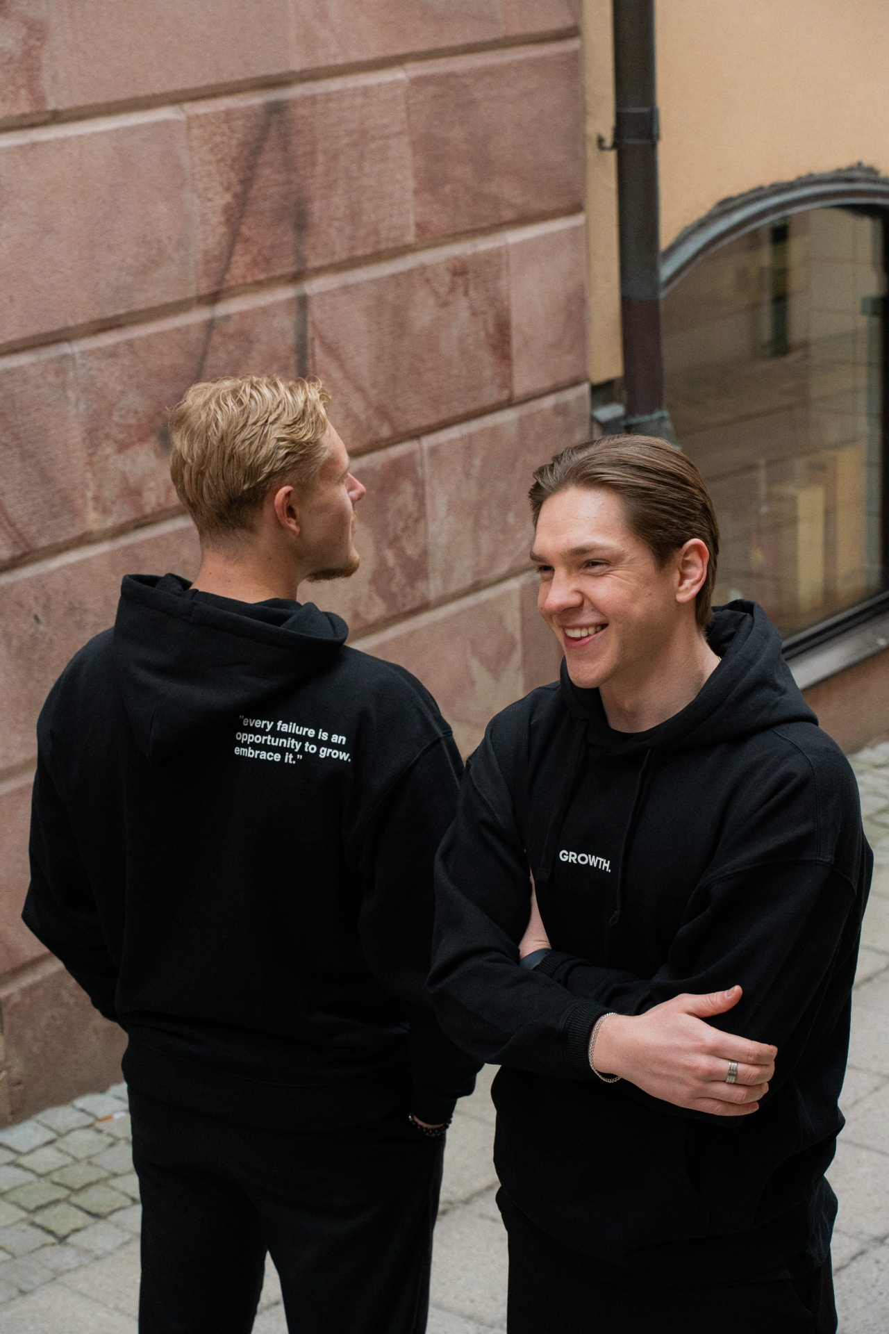 Two men in black GROWTH. hoodies on a Stockholm street, one showing the back print 'every failure is an opportunity to grow. embrace it.'